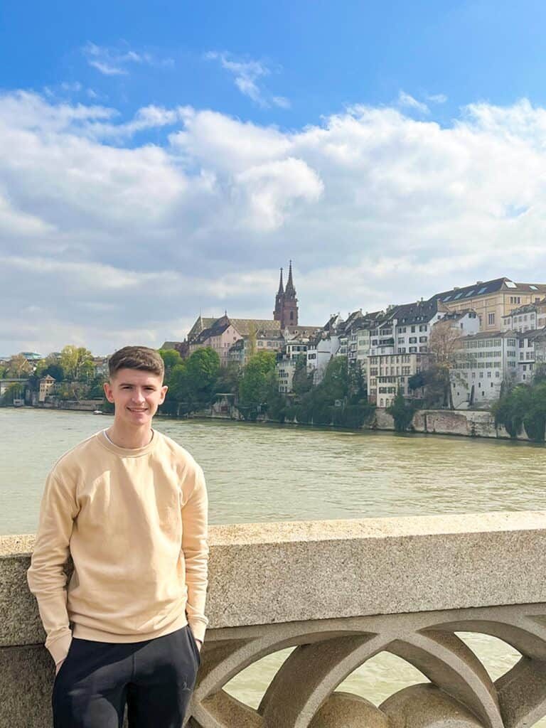 A young man standing by a balustrade overlooking the River Rhine with the Basel Minster and cityscape in the background, under a sky with scattered clouds.