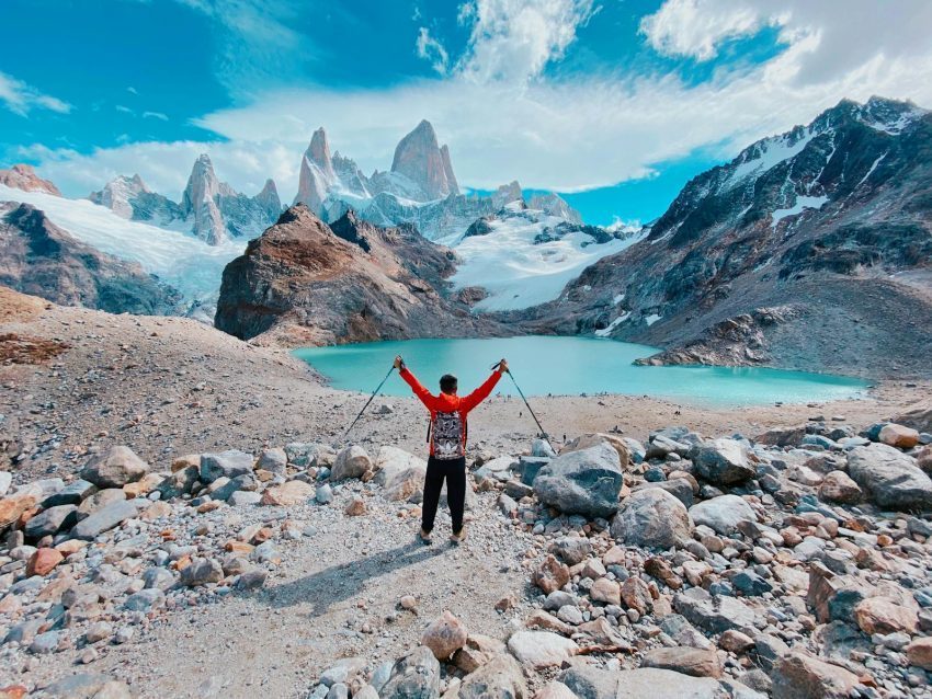 Man with arms raised in victory at Laguna de los Tres, Argentina, surrounded by stunning mountains and a turquoise lagoon.