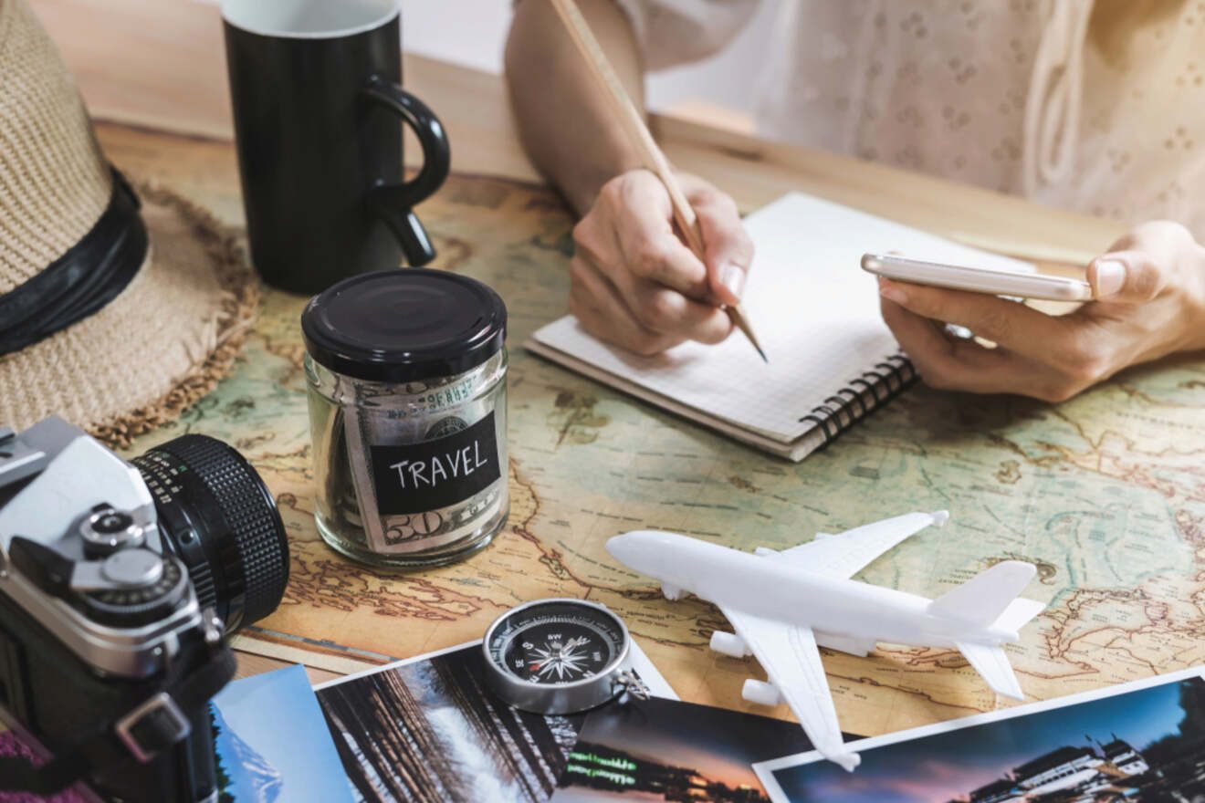 A person is planning a trip, writing in a notebook with travel items such as a map, camera, toy airplane, compass, and jar labeled 'Travel' on the table.