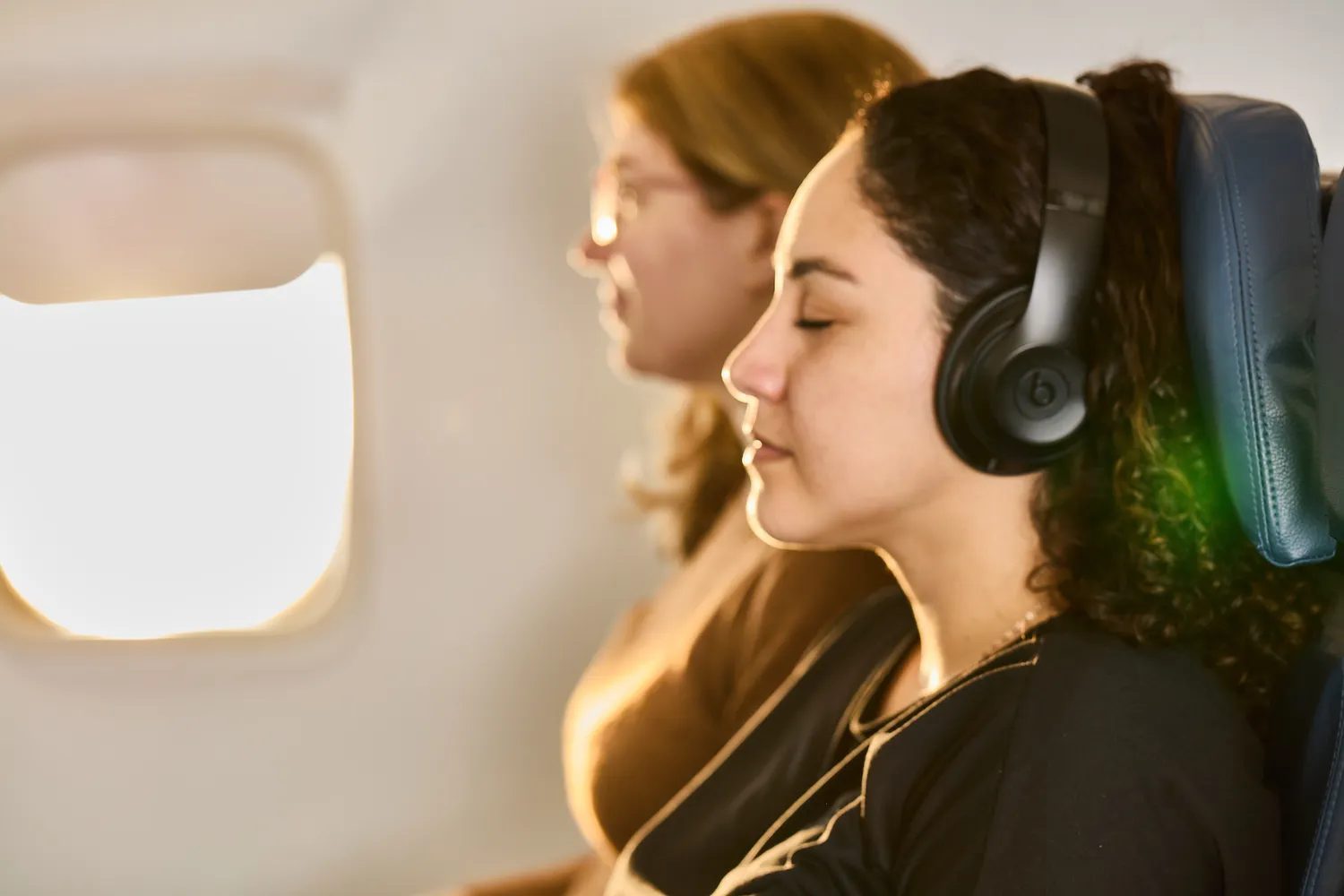 Two airplane passengers seated, one resting and one wearing headphones during a long flight