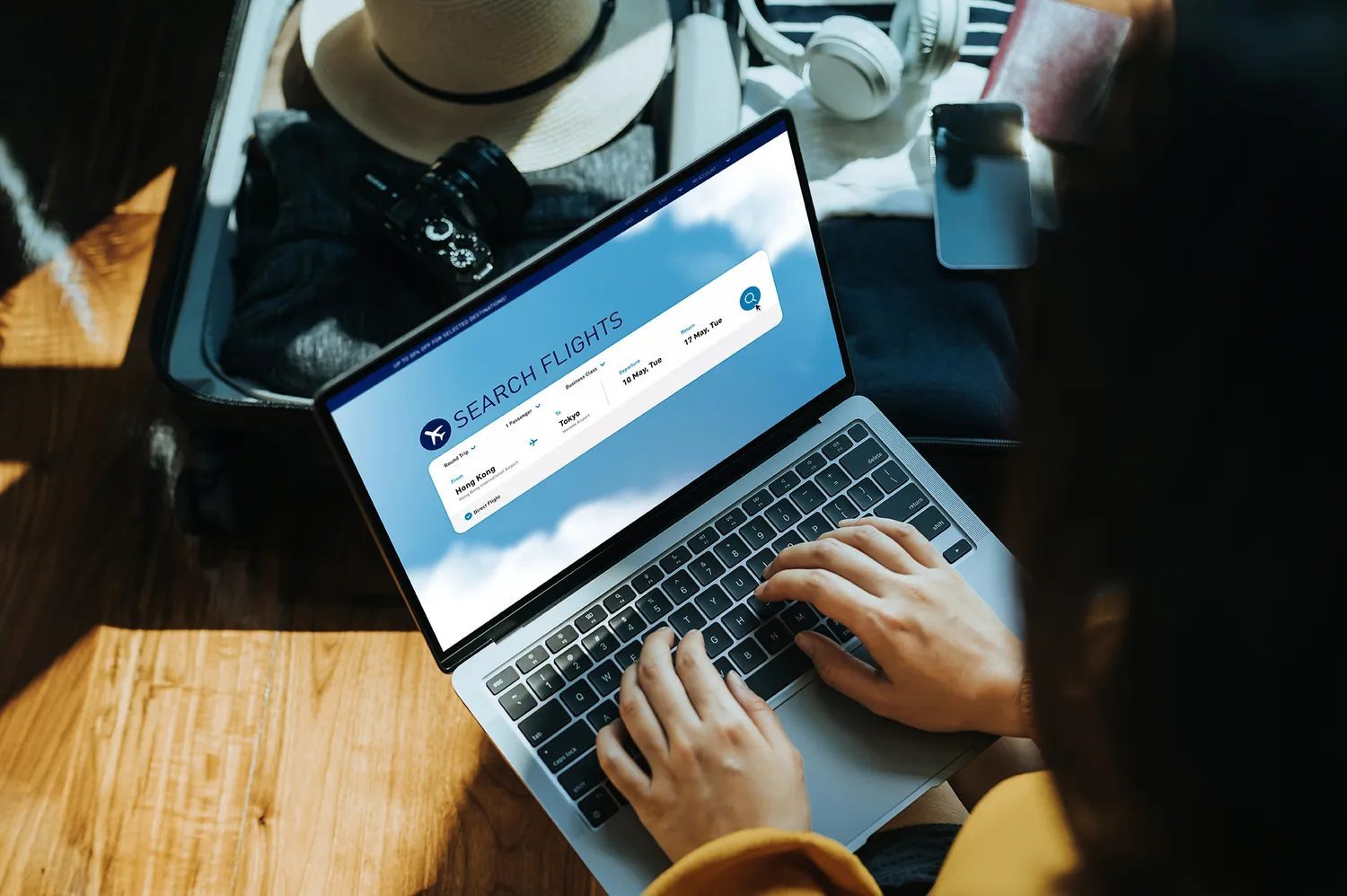 Person using a laptop to search for flights open suitcase and travel items in the background