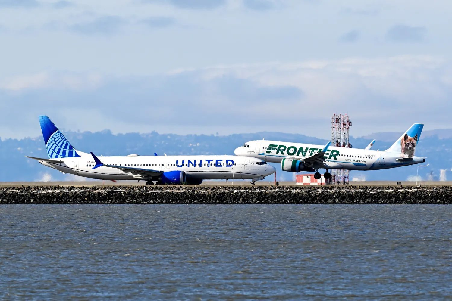 Two airplanes from United Airlines and Frontier Airlines parked on a tarmac near water one showing branding prominently