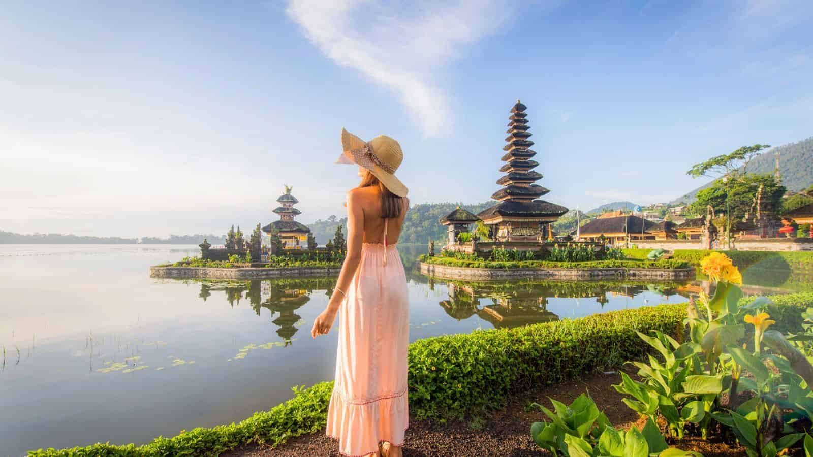 A woman in a long pink dress and wide-brimmed hat stands by a lake, looking at a traditional Balinese temple with tiered roofs, surrounded by greenery and water.