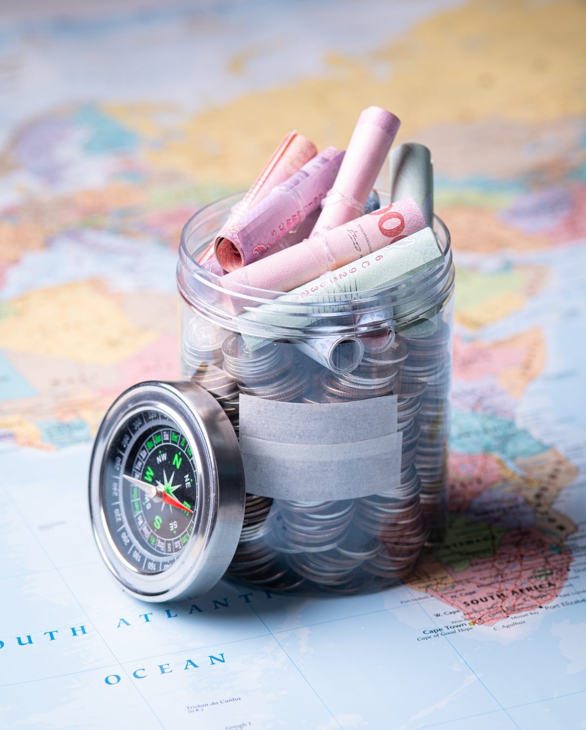 Close-up of a glass jar filled with money, a compass, and a passport on a world map, symbolizing travel savings and budgeting.
