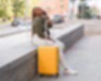 Woman with braided hair sits on a bench, talking on a phone, next to a bright yellow suitcase in an urban setting.
