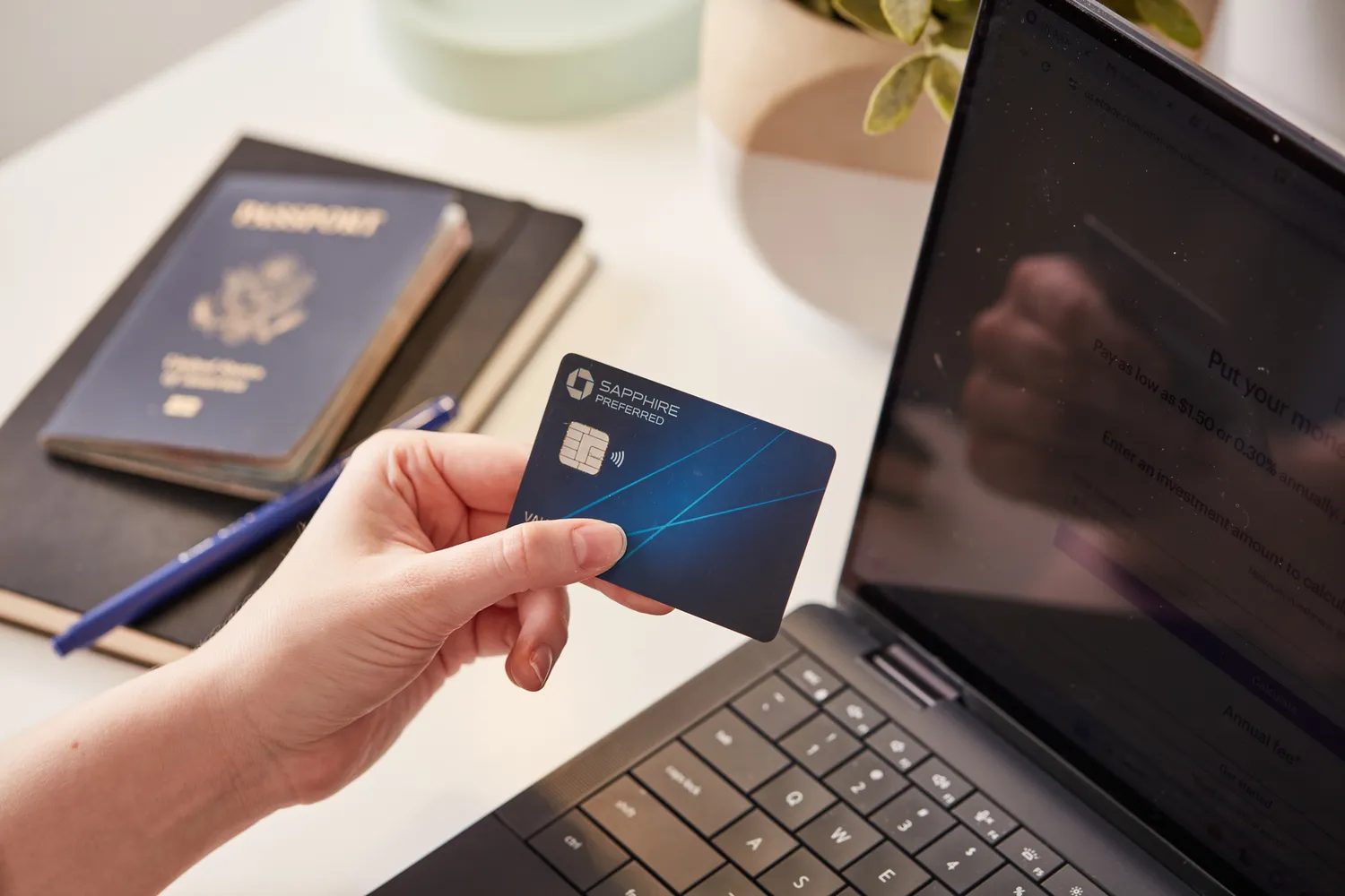 A hand holding a credit card in front of a laptop with a passport and notebook nearby