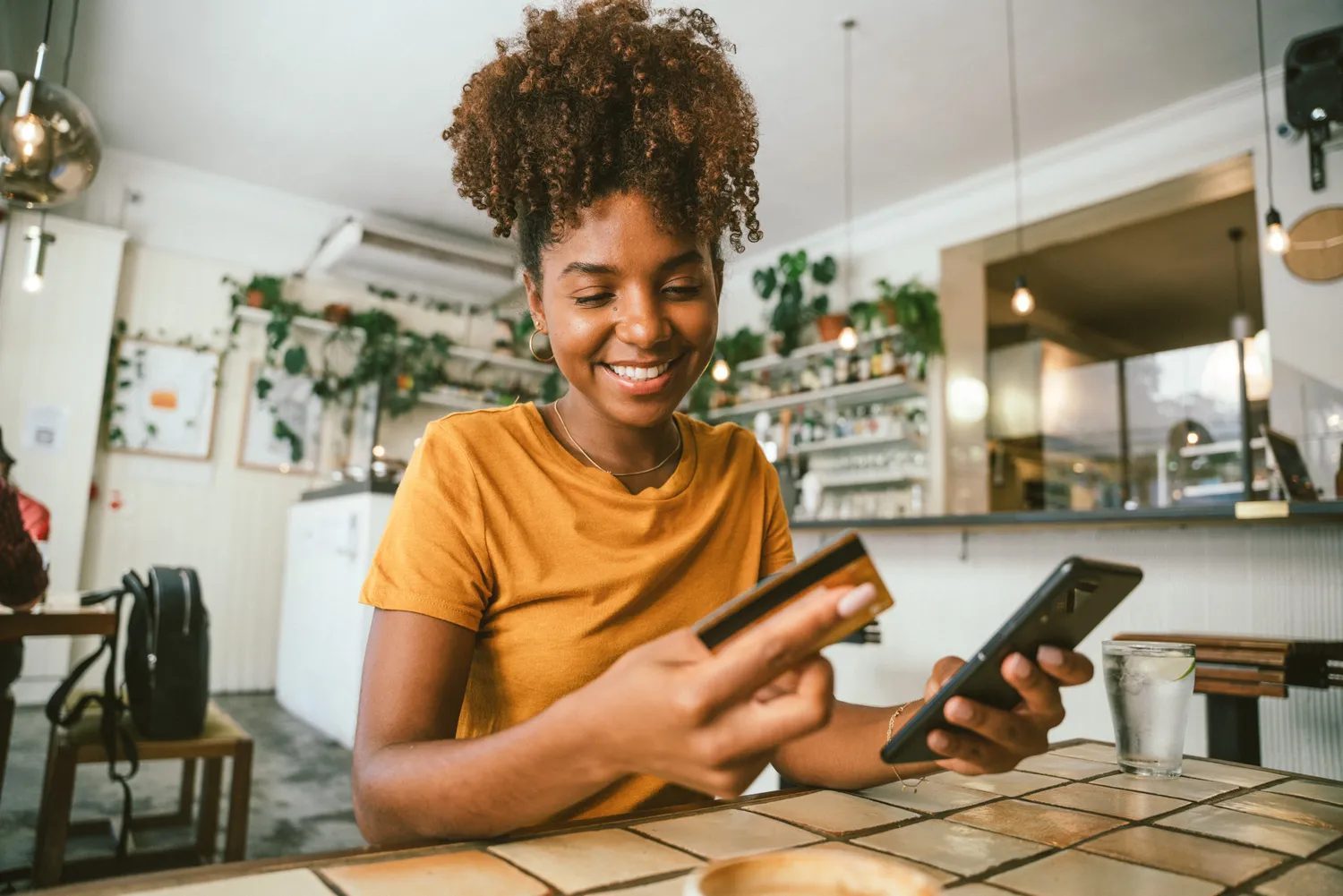A person sitting at a table in a cafe holding a credit card and a smartphone smiling
